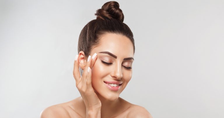 A woman with smooth, glowing skin and neatly groomed eyebrows smiles gently with her eyes closed. She has her dark hair styled in a high bun and lightly touches the side of her face with her hand, showcasing well-manicured white nails.