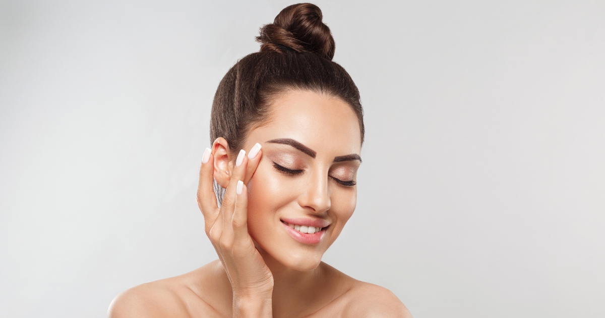 A woman with smooth, glowing skin and neatly groomed eyebrows smiles gently with her eyes closed. She has her dark hair styled in a high bun and lightly touches the side of her face with her hand, showcasing well-manicured white nails.