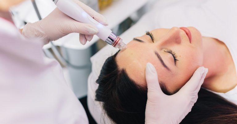A woman with dark hair lying comfortably with her eyes closed during a HydraFacial treatment. A skincare specialist in gloves uses a facial device on her forehead in a professional spa environment with white decor and soft lighting.