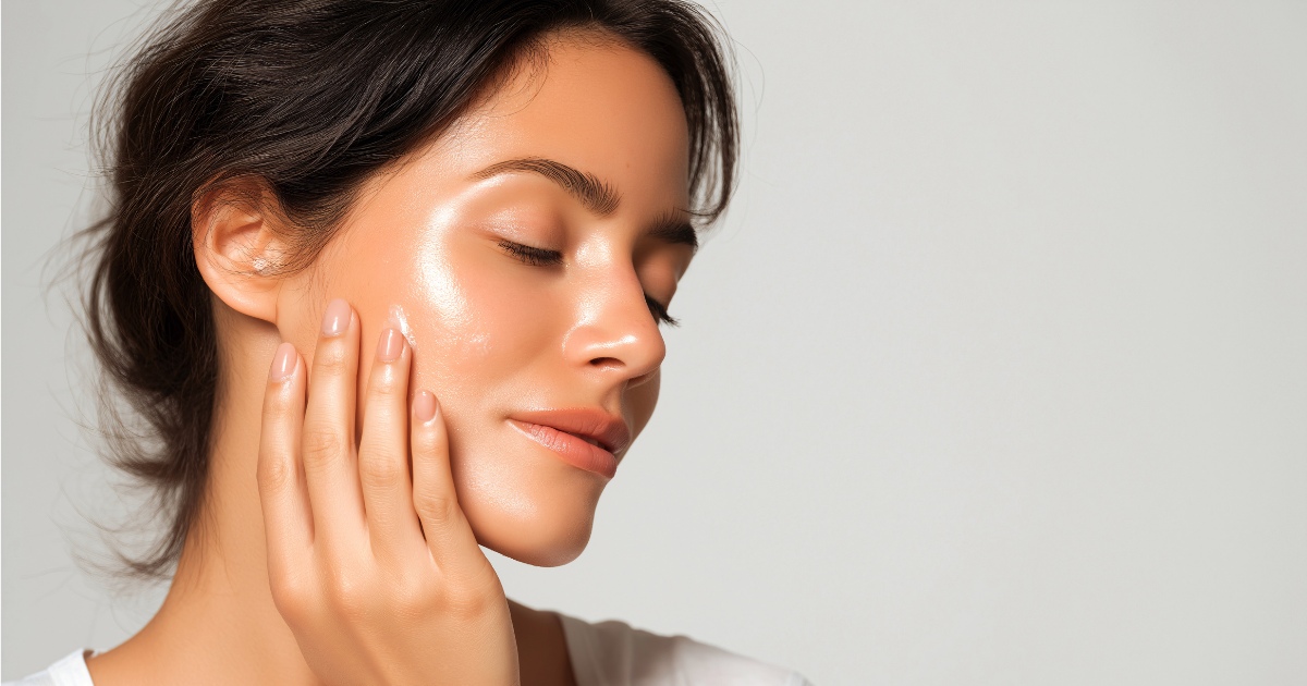 A woman with glowing, dewy skin rests her hand gently on her cheek, eyes closed and expression calm, showcasing smooth, radiant complexion against a soft, neutral background.