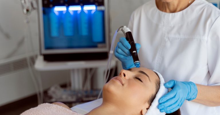 A person lying back with eyes closed receives a facial treatment while a clinician wearing blue gloves uses a handheld skincare device on their forehead.