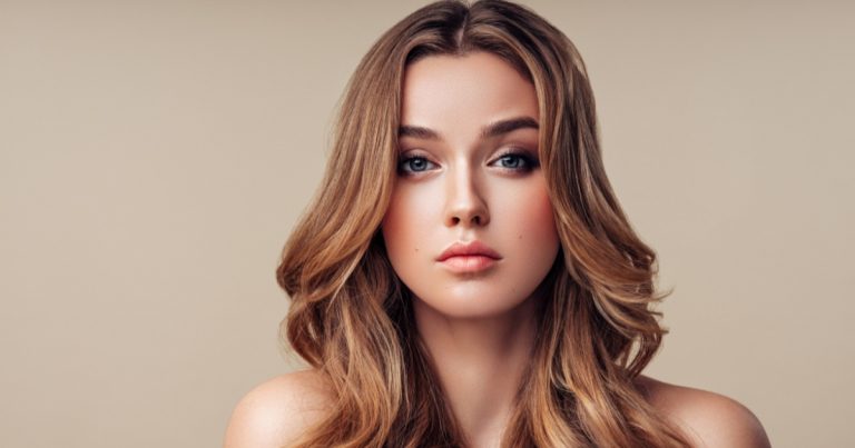 Close-up portrait of a young woman with long, wavy light brown hair and blue eyes, facing forward against a neutral beige background, with soft natural makeup and a calm, neutral expression.